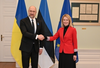 Ukraina peaminister tumedas ülikonnas (vasakul) ja Eesti peaminister punases jakis ja tumedas seelikus (paremal) lippude ees kätlemas. Taustal on Ukraina, Eesti ja Euroopa Liidu lipud